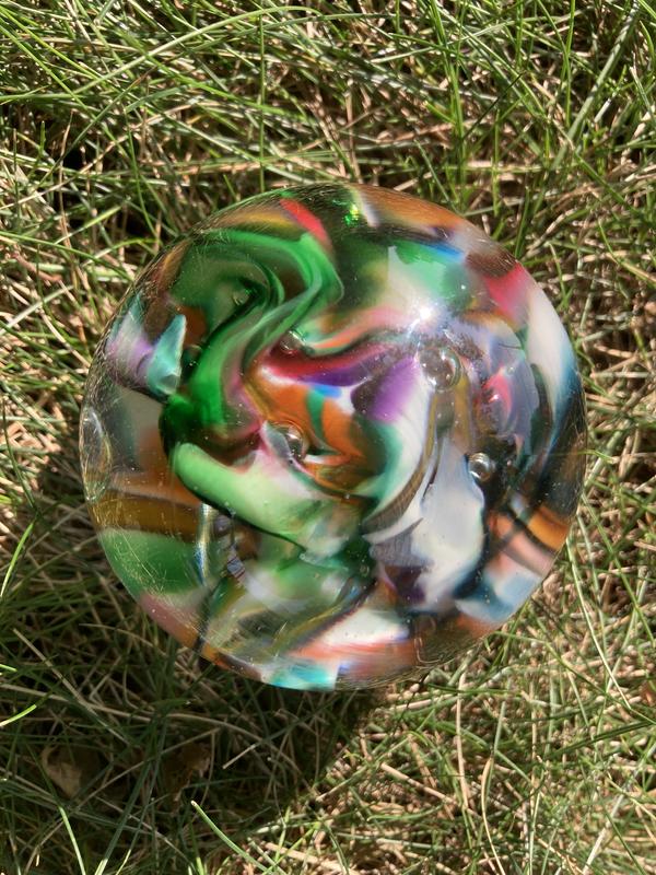 A photograph of a glass globe resting on green and brown grass. The globe contains a riot of swirling colors, including dark and light green, white, burgundy, dark blue, orange and purple.