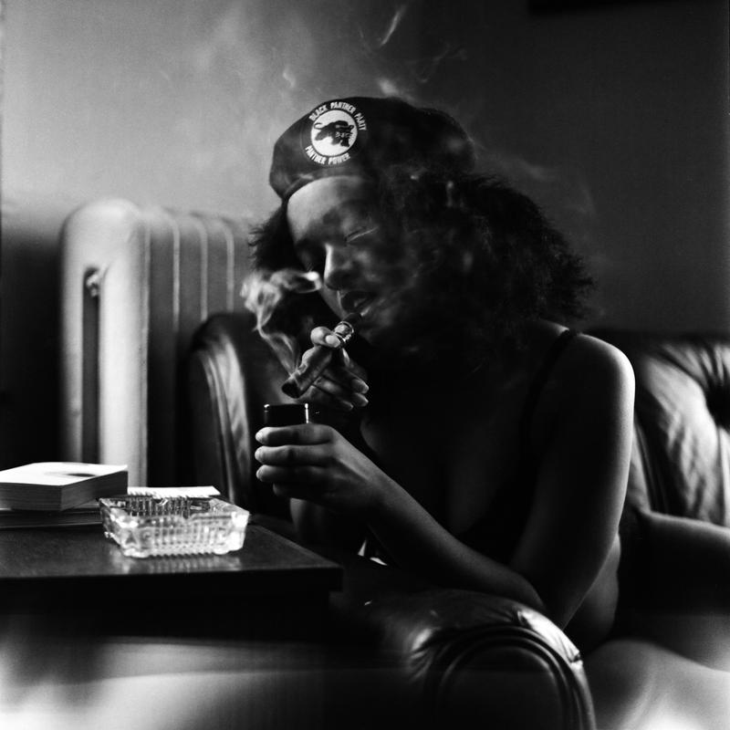The image depicts a black and white photograph of a woman smoking a cigar on a couch wearing a Black Panther Berret.