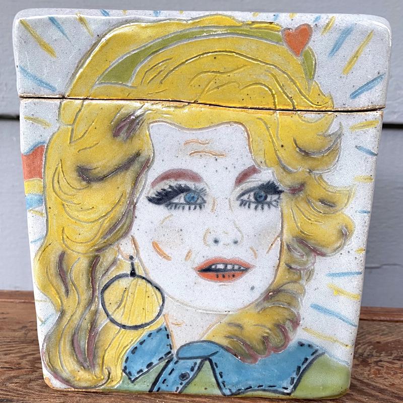 A Ceramic Box with an etched drawing of Dolly Parton. The top has the name "Dolly" etched in cursive. The other 3 sides feature a guitar, rainbows and flowers