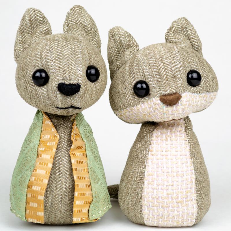 Two small stuffed animals made of neutrally-toned wool sit together. One wears a green silk cloak with orange trim.