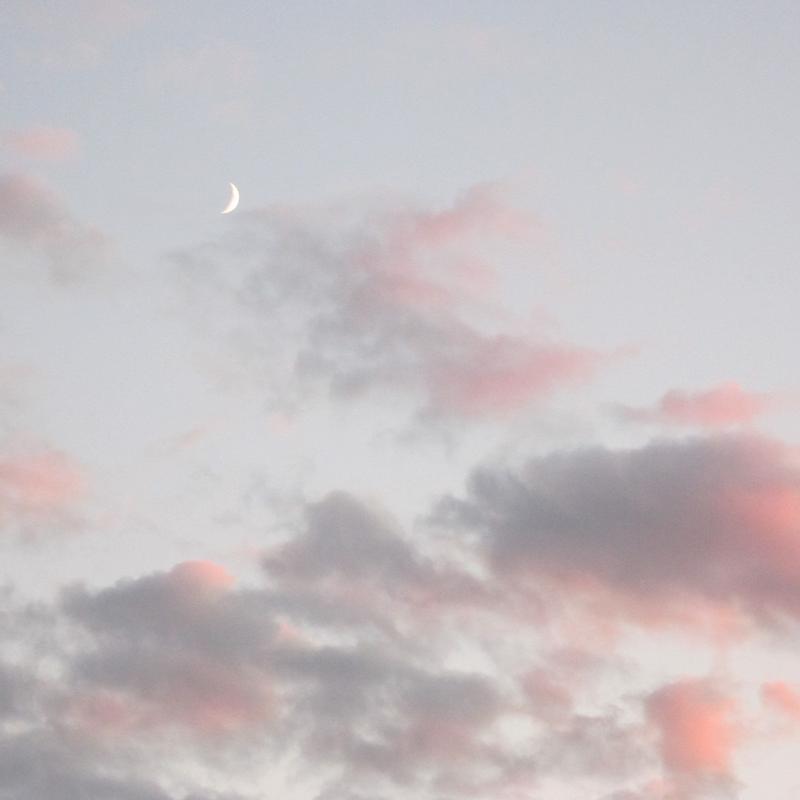 A photograph of a waxing crescent moon against a blue-gray sky with pink and gray clouds.