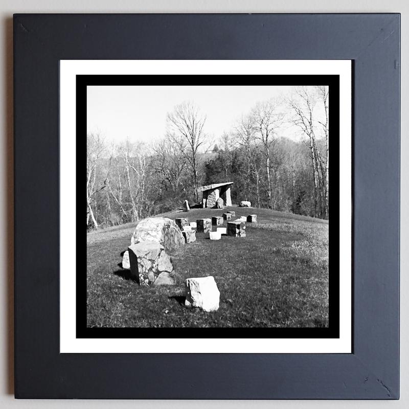 A black and white photograph of a group of large stones placed in the ground in a curved line, reminiscent of a slithering snake, with woods in the background.