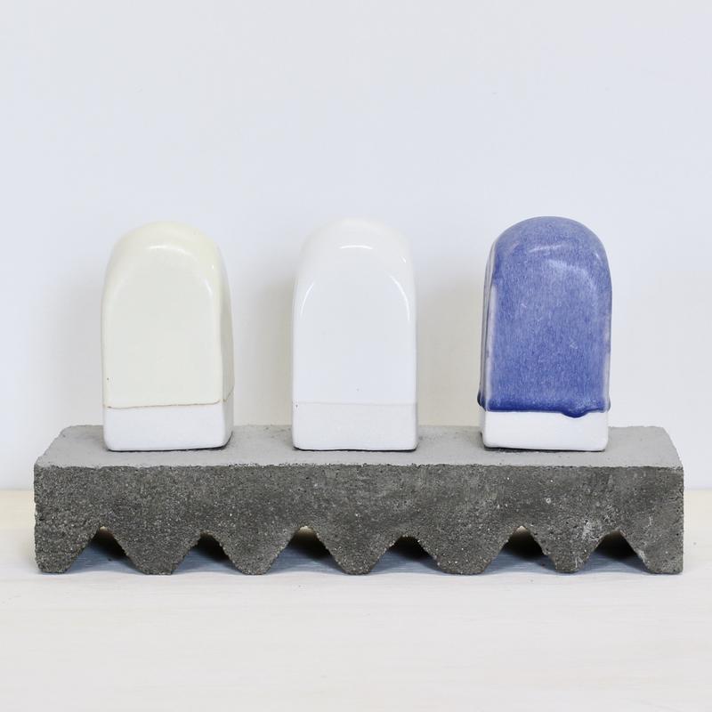 A sculpture with a cement base having a zig zag foot. It is holding up three arched shaped ceramic forms that are off white, white, and blue.
