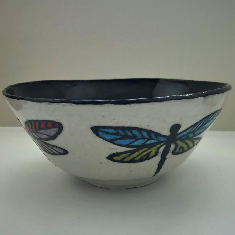 A sgraffito style white clay bow with a black inner surface. Vertical dragonflies adorn the outside of the bowl with colorful wings in blue and green and purple and red.